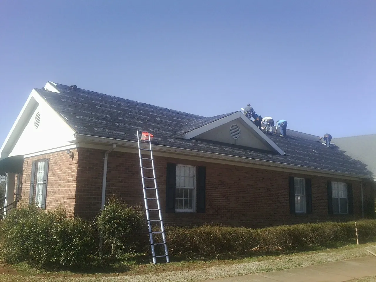 Crew tearing shingles off a brick ranch roof with a ladder set — Emergency Roof Repair work by our crew in Ely