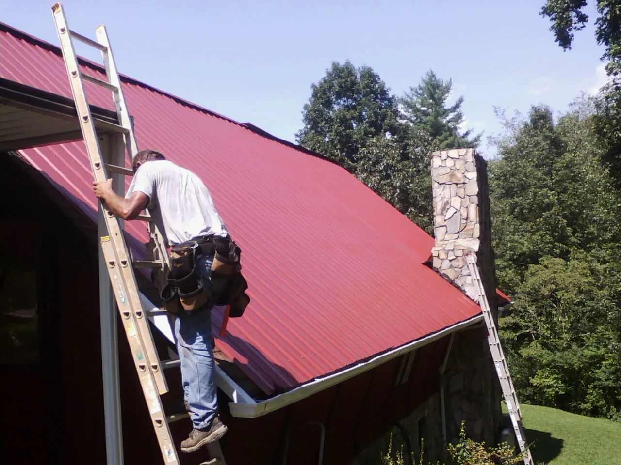 Roofer on a ladder beside a red metal roof and stone chimney — Soffit & Fascia Repair by Roofing Brothers in Ely